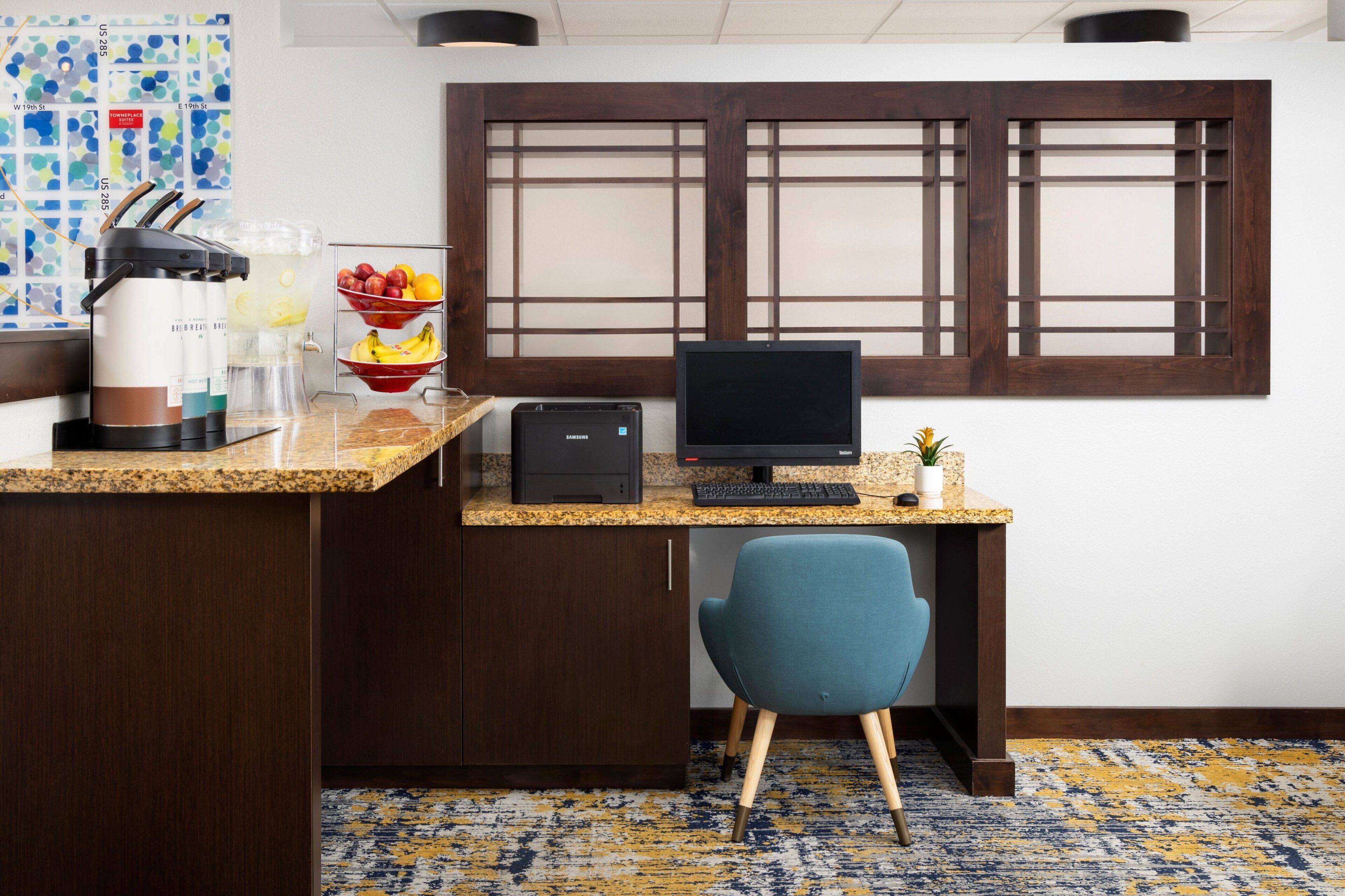 A hotel room with a kitchenette, a desk with a computer, a blue chair, a window with wooden blinds, and a patterned carpet.