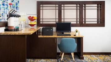 A hotel room with a kitchenette, a desk with a computer, a blue chair, a window with wooden blinds, and a patterned carpet.