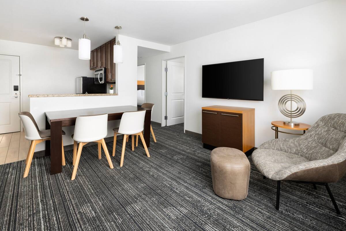 A modern hotel room with a dining area, a flat-screen TV, a lamp, and a sofa.