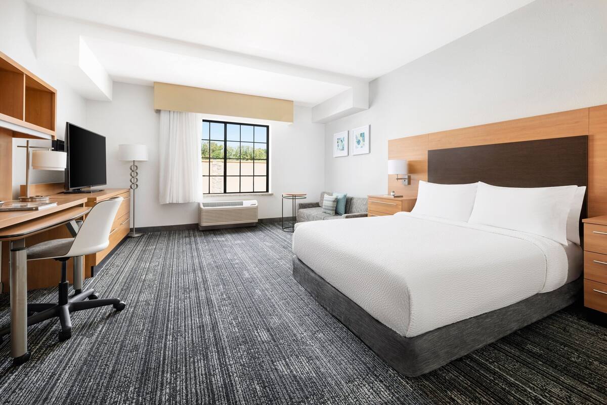 A hotel room with a bed, desk, chair, television, and a window with a view of greenery.