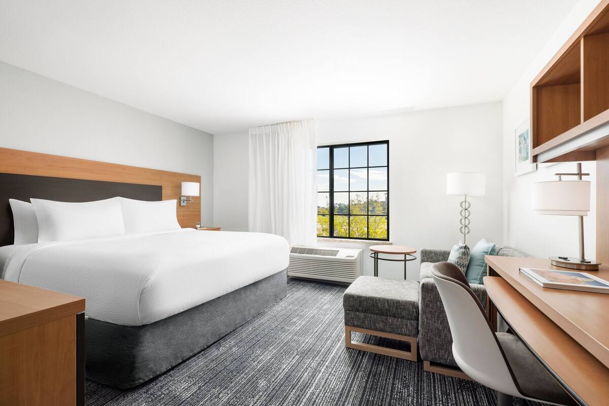 A hotel room with a large bed, a sofa, a desk, and a window with a view of trees.
