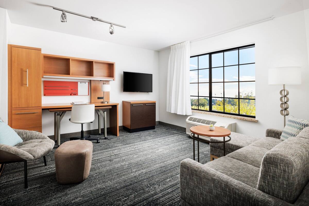 A modern hotel room with a kitchenette, a flat-screen TV, a large window with a city view, and a cozy seating area.