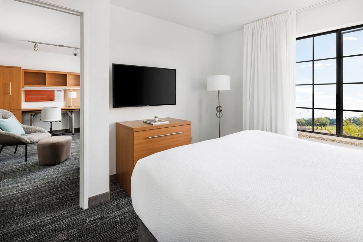 A hotel room with a large bed, a flat-screen TV, a wooden desk, a chair, and a view of the outdoors through a large window.
