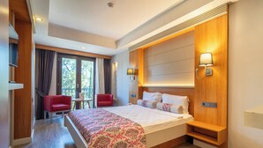 A hotel room with a bed, a red chair, a small table, a window with curtains, and a wall-mounted lamp. - May Park Hotel (Izmir)