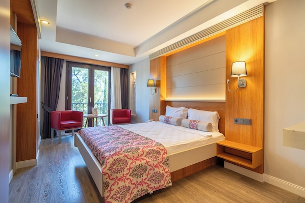 A hotel room with a bed, a red chair, a small table, a window with curtains, and a wall-mounted lamp.
