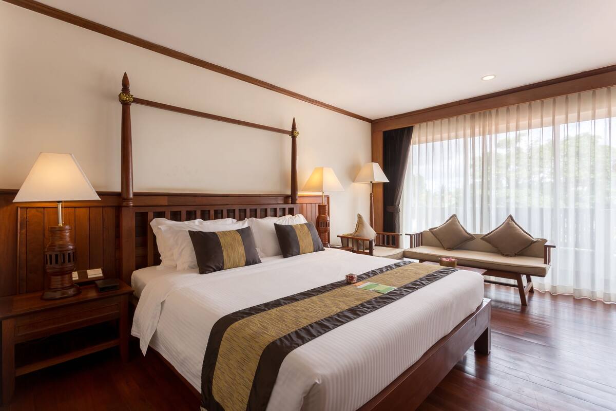 A spacious hotel room with a large bed, wooden headboard, two bedside tables with lamps, a bench with cushions, and a large window with sheer curtains.