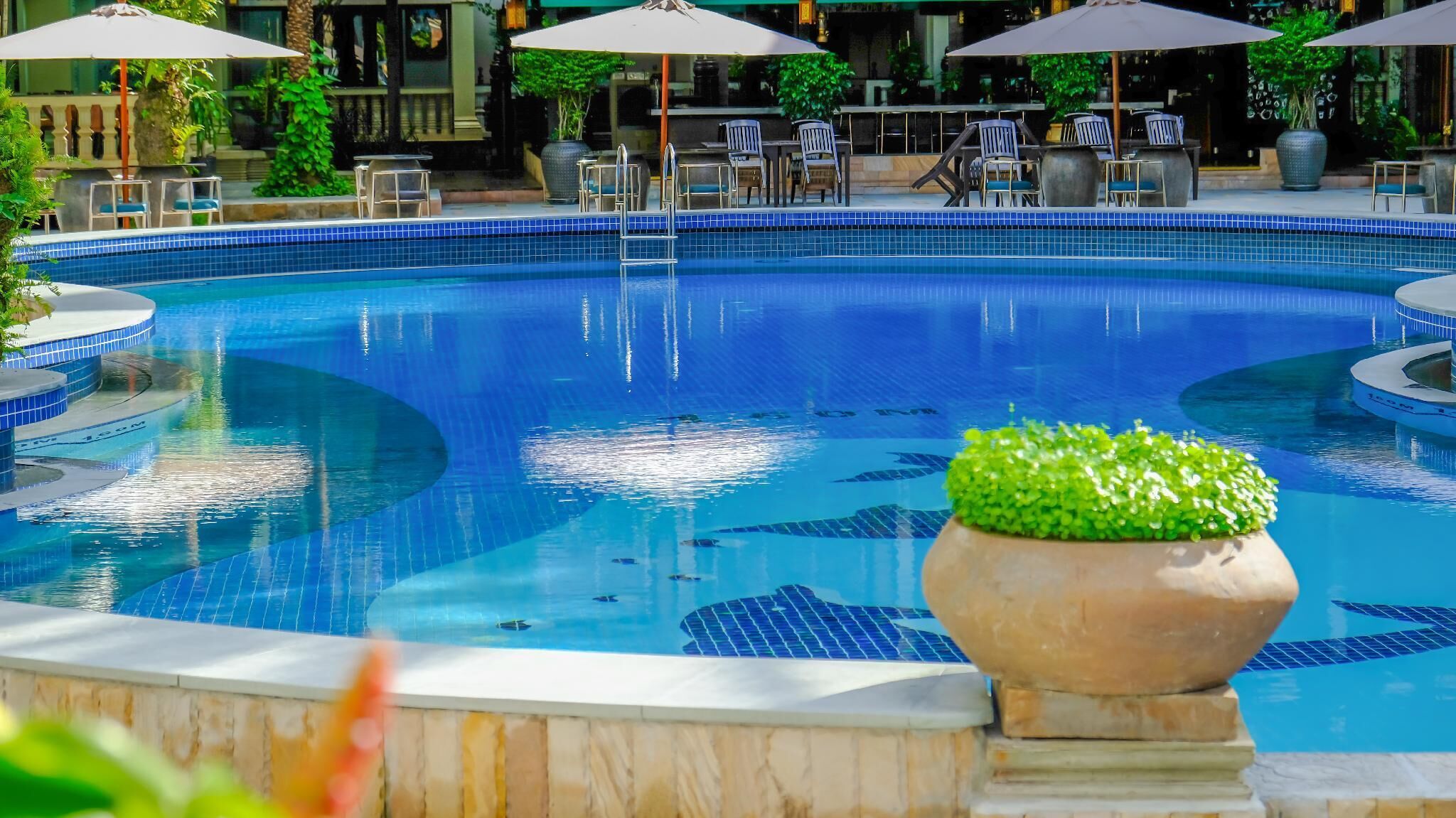 Una piscina grande y cristalina de color azul con borde de azulejos, rodeada de sillones de descanso y sombrillas.