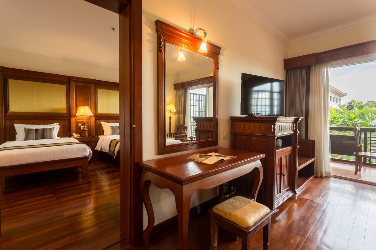 A hotel room with two beds, a wooden desk, a television, and a balcony with a view of greenery.