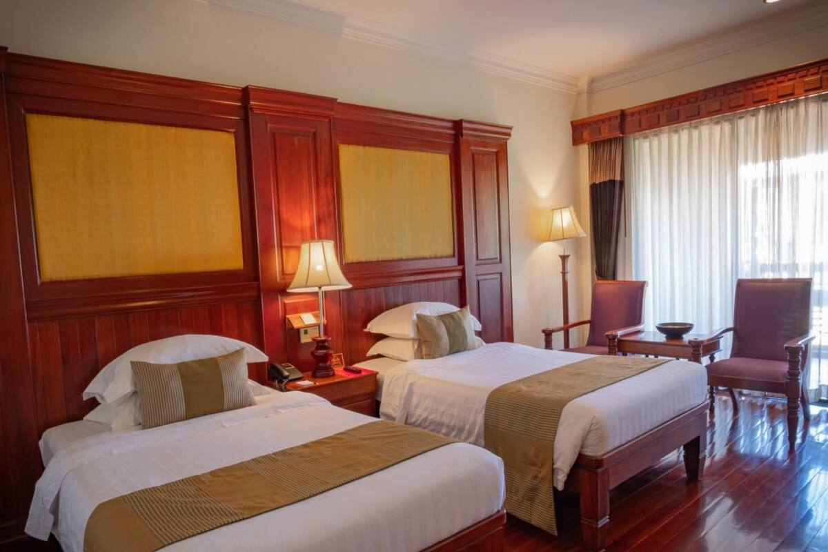 A hotel room with two beds, wooden paneling, a desk with a chair, and a large window with curtains.