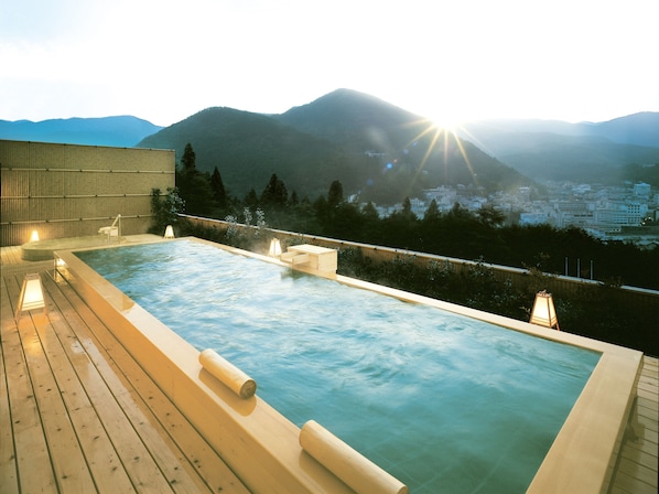 A rooftop infinity pool with wooden deck and lounge chairs, overlooking a mountainous landscape.