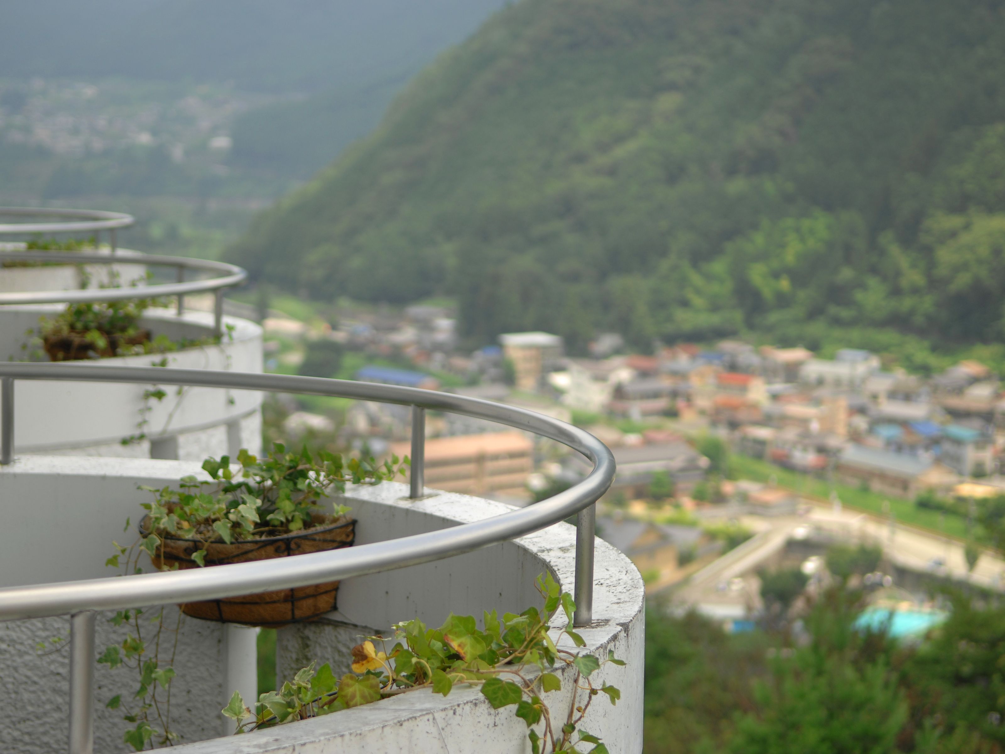 Un balcon doté d’une balustrade en métal, offrant une vue sur le paysage urbain et une région montagneuse.
