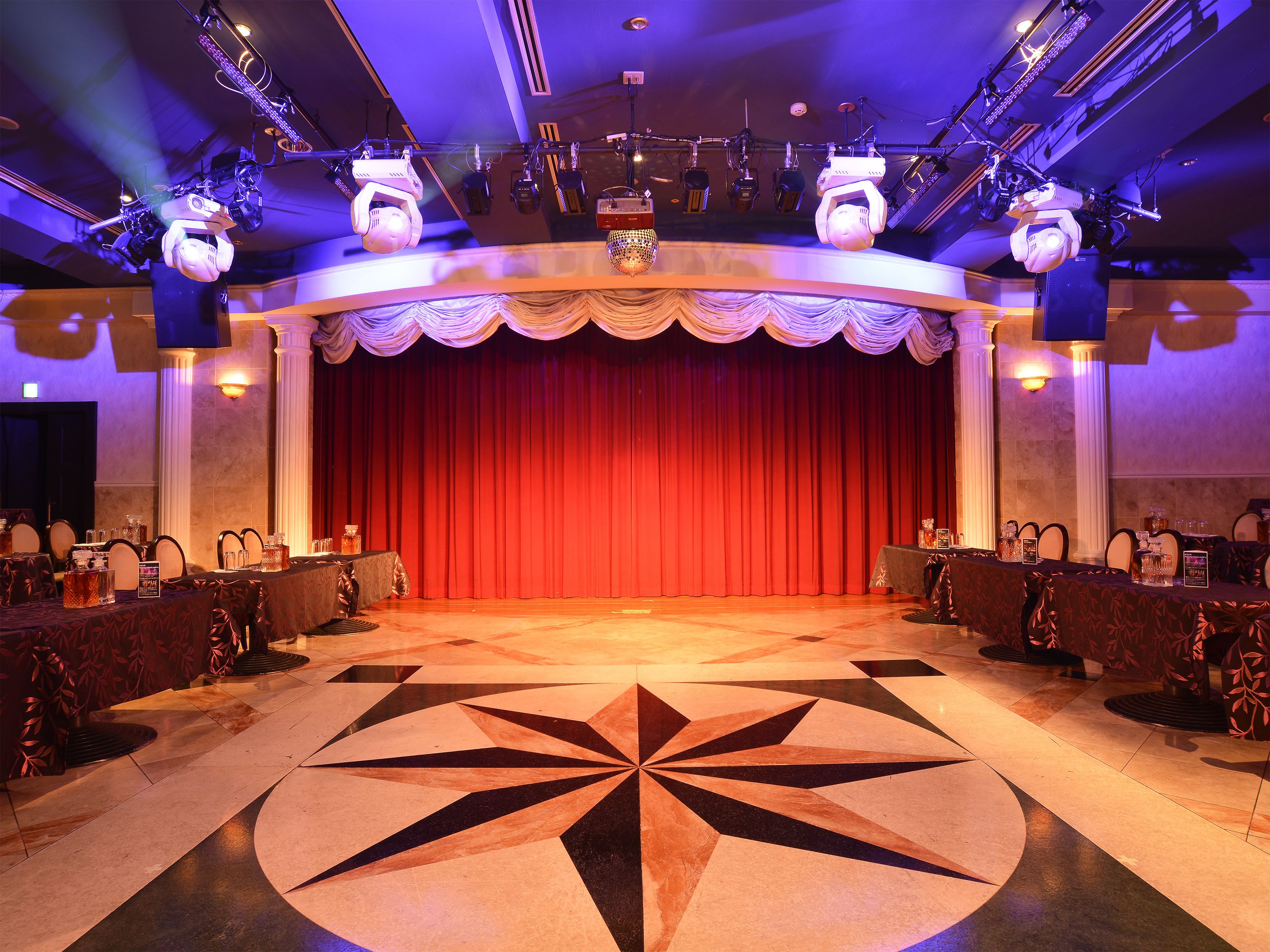 A stage with a red curtain, a patterned floor, and tables with chairs arranged for an event.