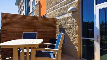 Een terras buiten met een houten tafel en stoelen, een stenen muur en een glazen deur die leidt naar een balkon.