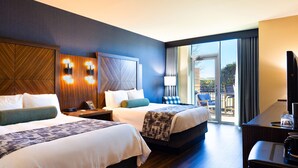 Habitación de hotel con dos camas, un escritorio, un televisor y un balcón con mobiliario exterior.
