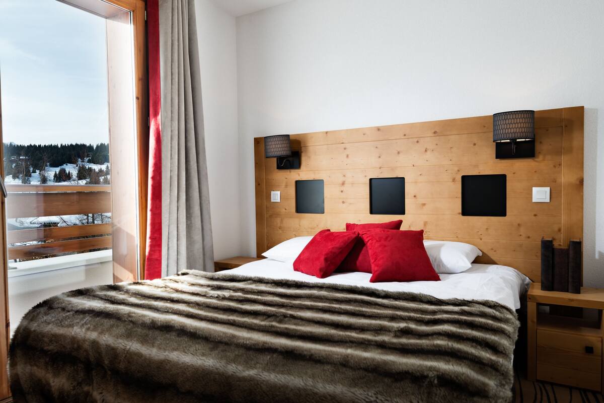 Habitación de hotel con cabecera de madera, un ventanal con vistas a un paisaje nevado, una manta de piel sobre la cama y dos almohadas rojas.