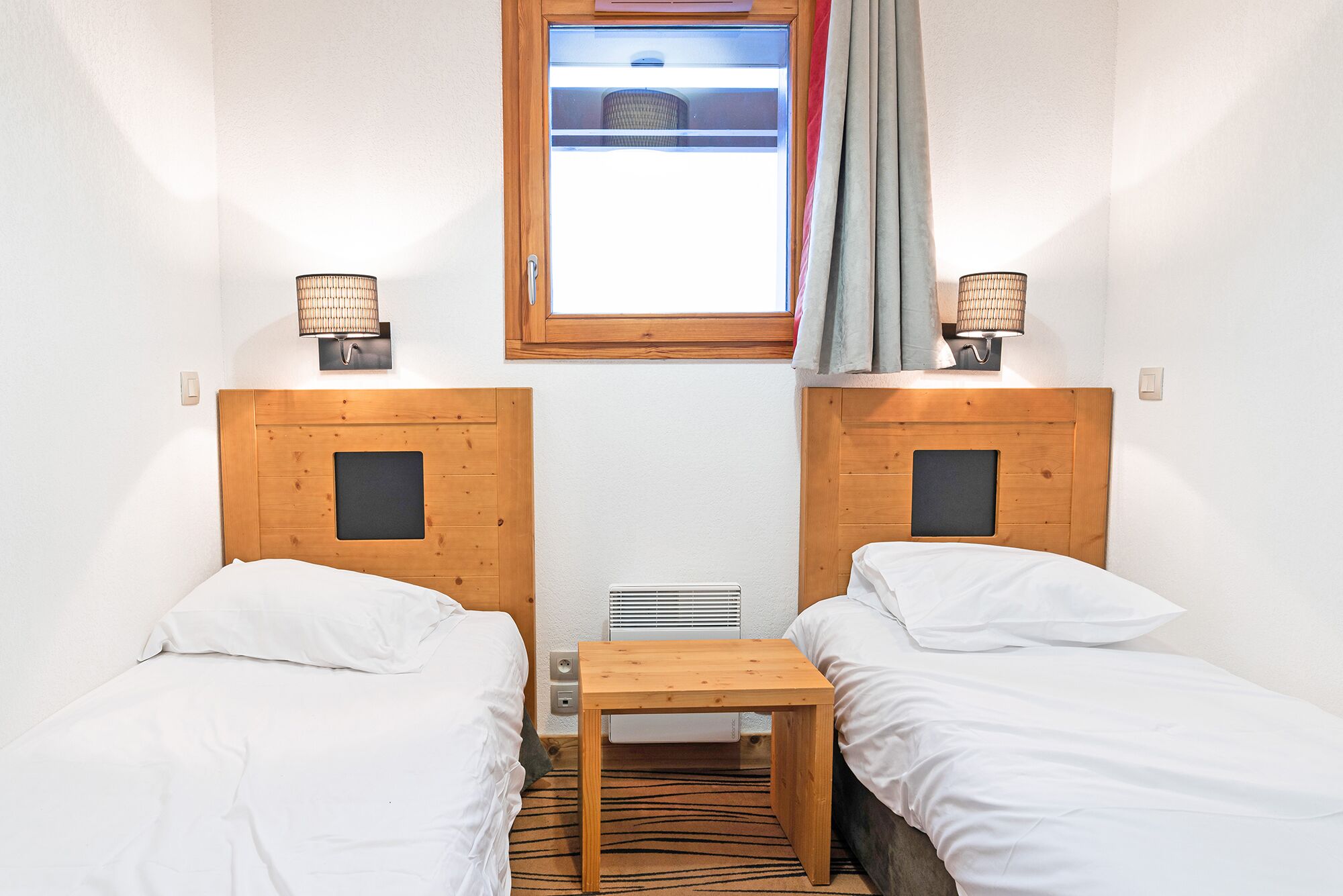A small, well-lit room with two single beds, a small table, and a window with curtains.