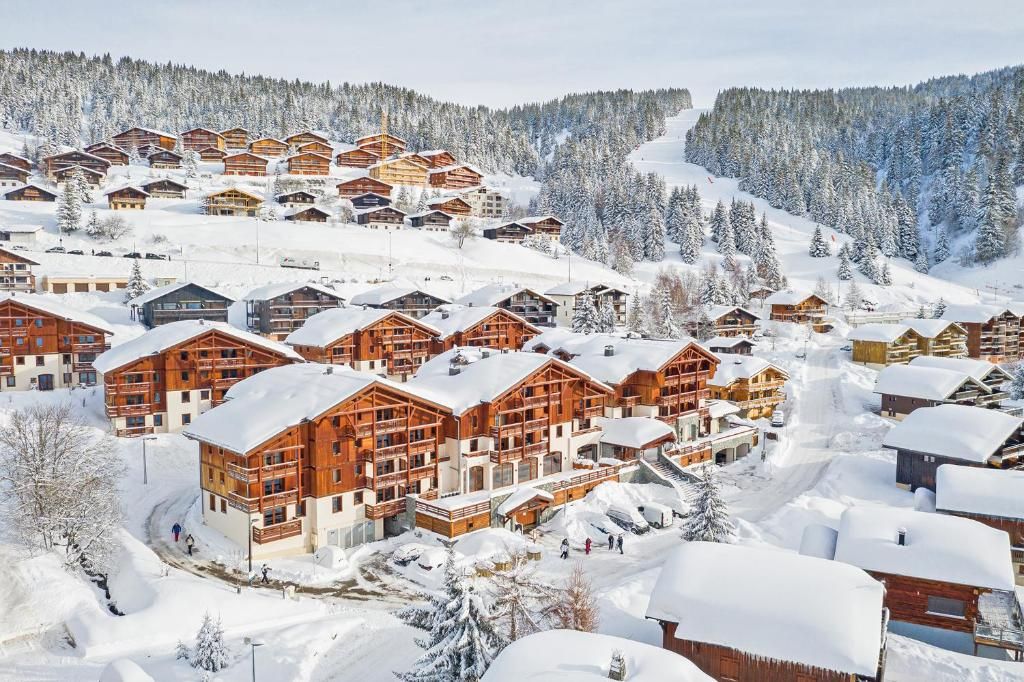 A snow-covered resort with multiple wooden lodges, a ski slope in the background, and a clear sky.
