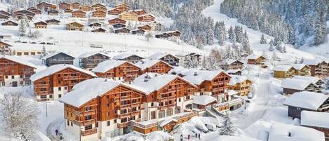 A snow-covered resort with multiple wooden lodges, a ski slope in the background, and a clear sky.