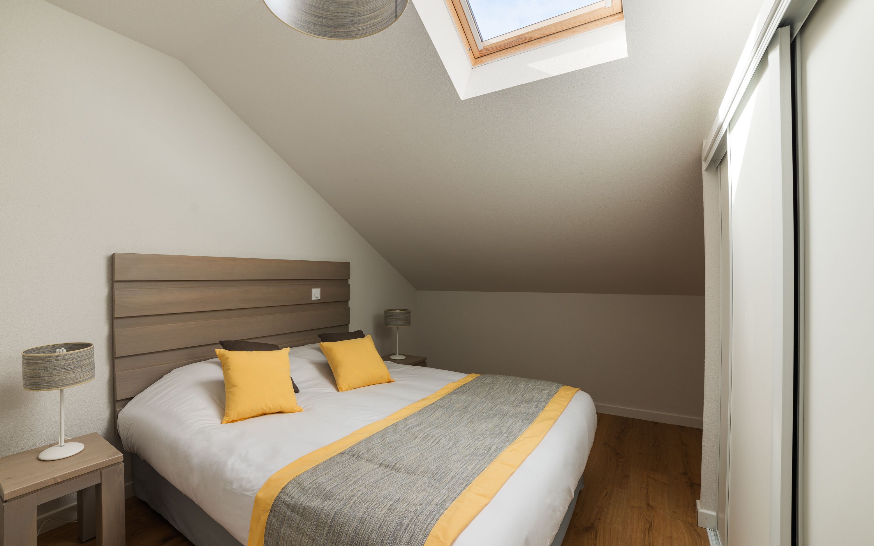 A bedroom with a sloped ceiling, a bed with a wooden headboard, two bedside tables, a lamp, and a window with a view.
