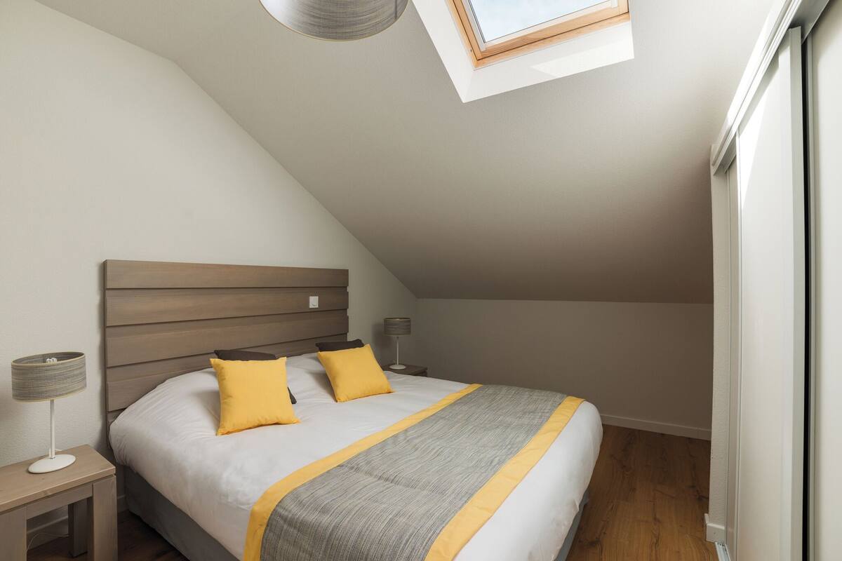 Een slaapkamer met een schuine plafond, een dakraam, een bed met hoofdeinde, twee nachtkastjes met lampen en een raam.