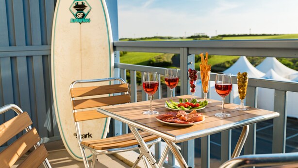 Una mesa con una tabla de surf, copas de vino y aperitivos en un balcón con vistas.