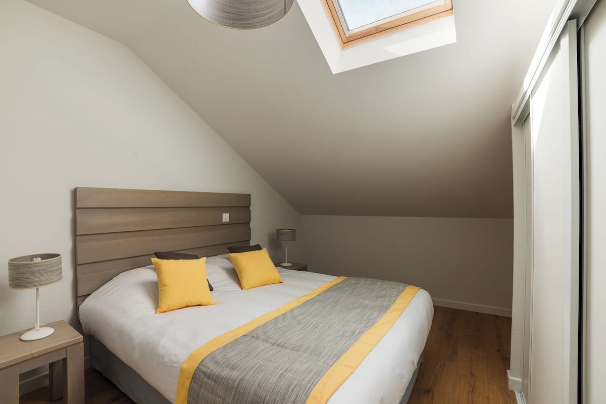 A bedroom with a sloped ceiling, a skylight, a bed with a headboard, two bedside tables with lamps, and a window.