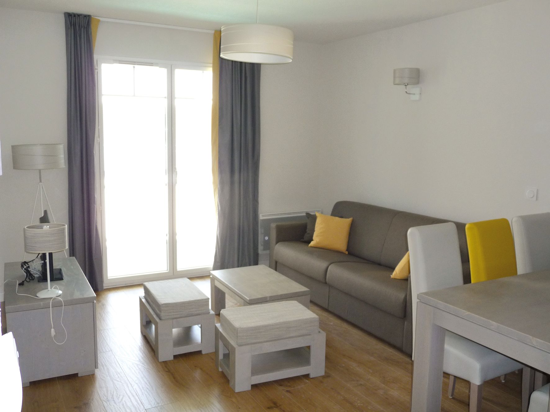 Un salon moderne avec un canapé gris, deux poufs blancs, une petite table et une lampe.