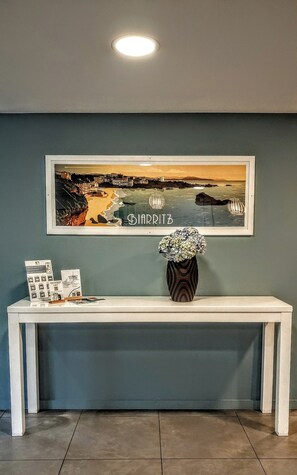 En inramad tavla som förestÀller en kuststad, en vit konsolbord med en vas med blommor och en inramad informationsbroschyr.