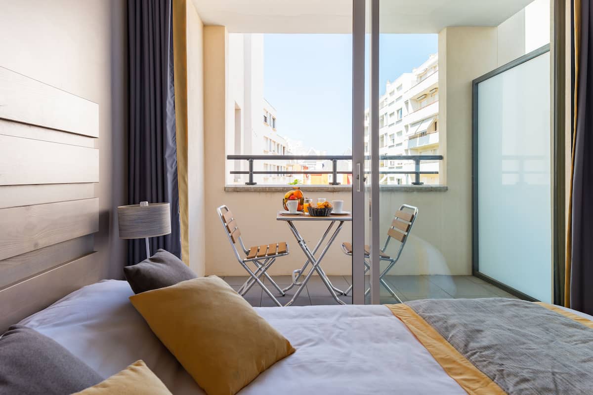 Una habitación de hotel moderna con cama, una mesita con desayuno, una silla y un balcón con vistas a edificios.