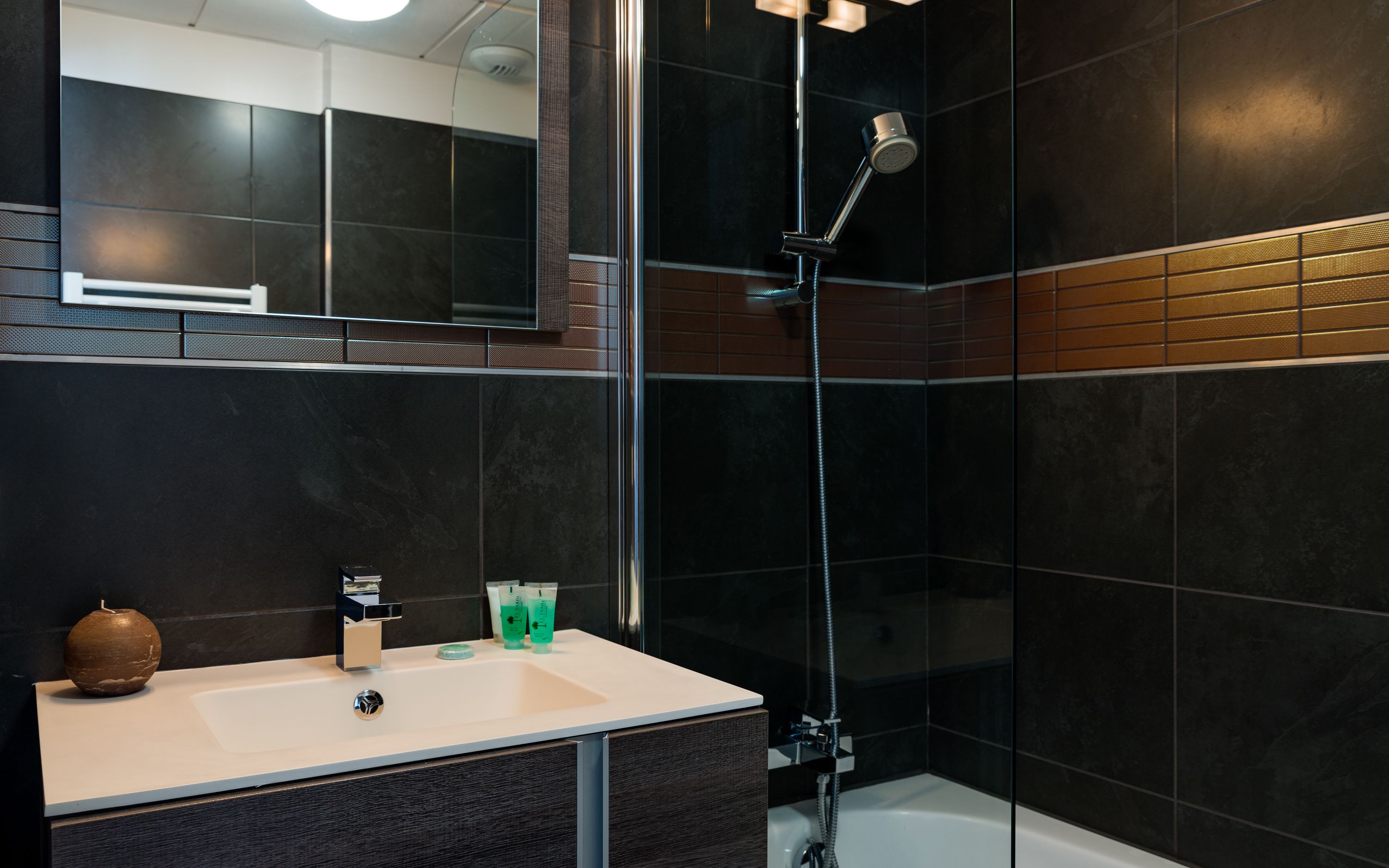 A modern bathroom with a dark tile wall, a white sink, and a shower with a handheld showerhead.