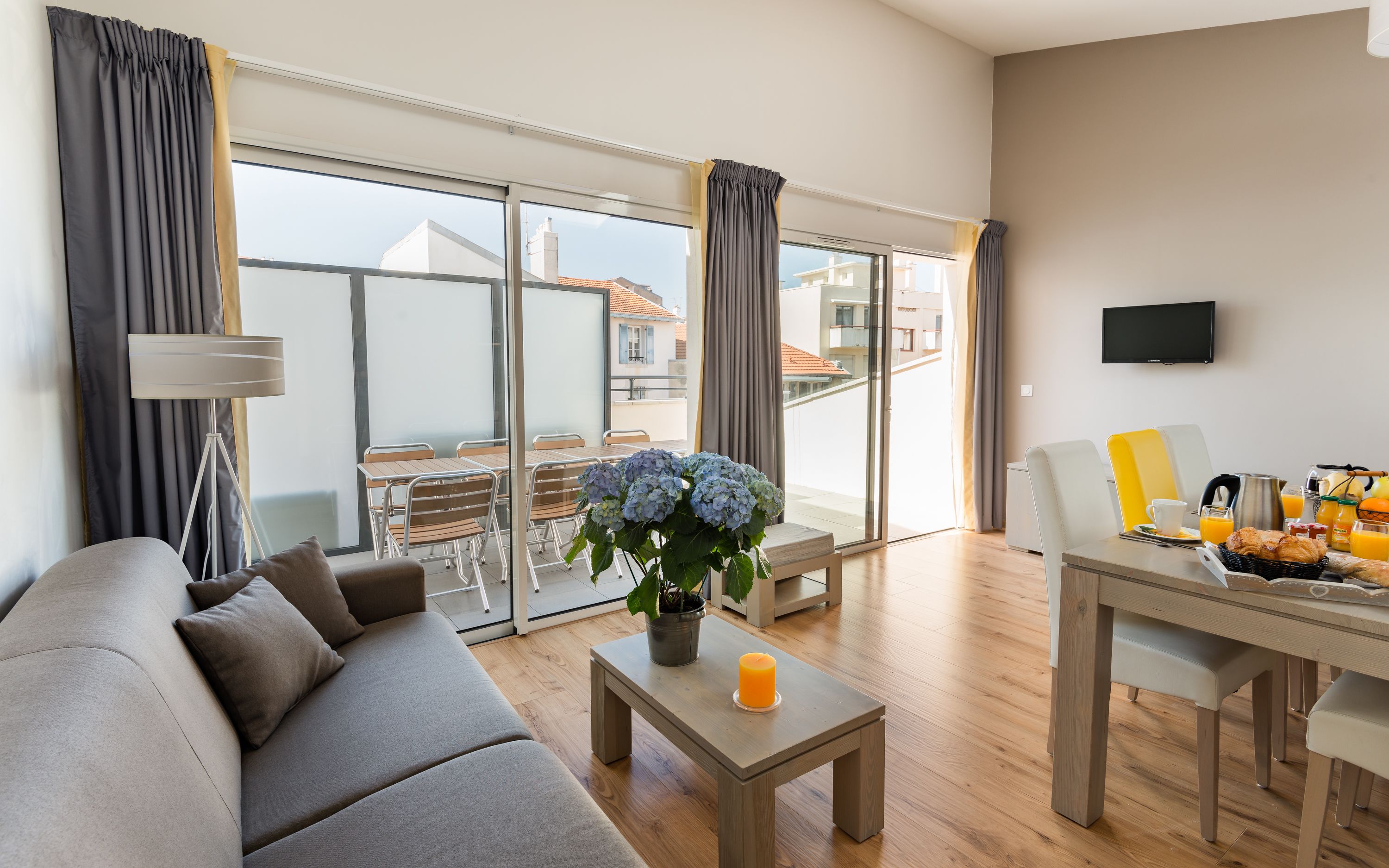 A modern living room with a sofa, a coffee table with a candle, a dining area with breakfast items, and a balcony with curtains.