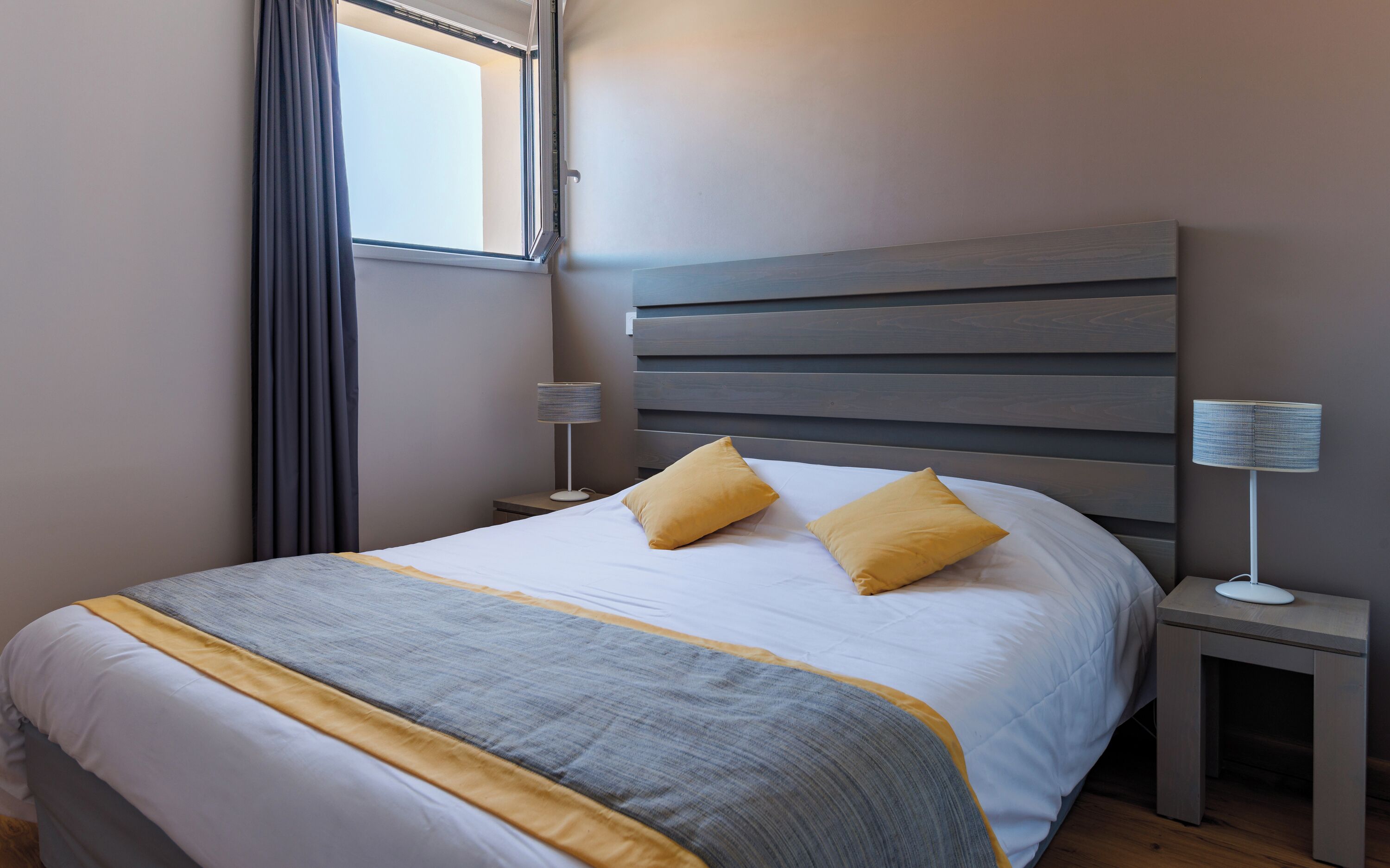 A neatly made bed with a grey and yellow bedspread, a wooden headboard, a bedside lamp, and a window with curtains.