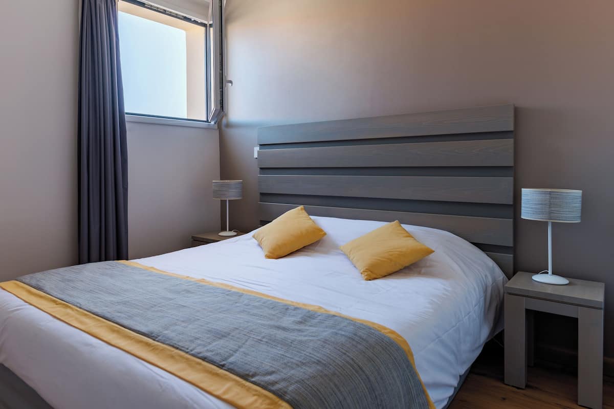 Una cama bien tendida con colcha gris y amarilla, cabecero de madera, lámpara de noche y una ventana con cortinas.
