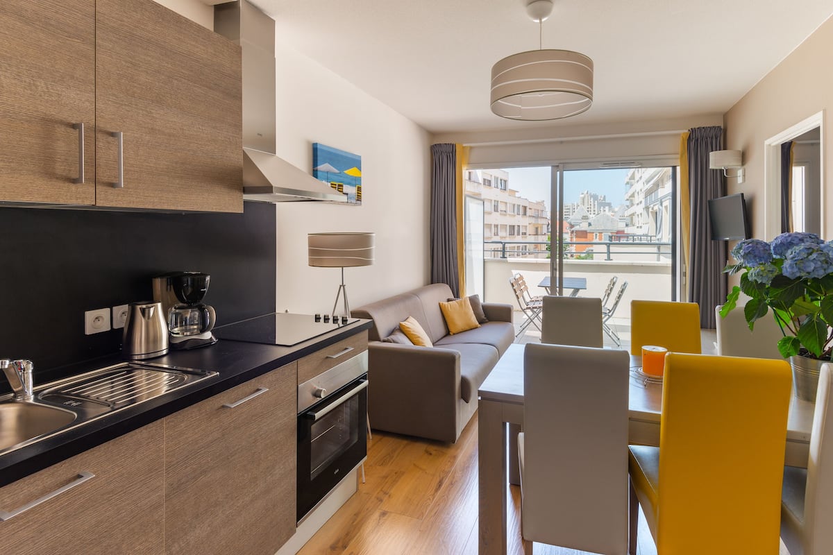 Una cocina moderna con cafetera, horno y lavaplatos. Hay una zona de comedor con mesa y sillas, y un sofá.