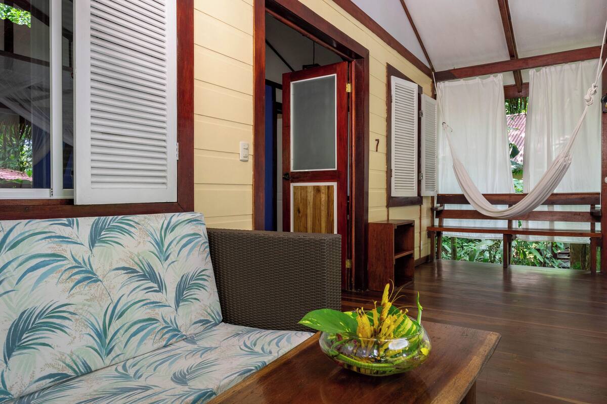 A room with a sofa, a wooden table with a bowl of fruit, a hammock, and a door leading to another room.