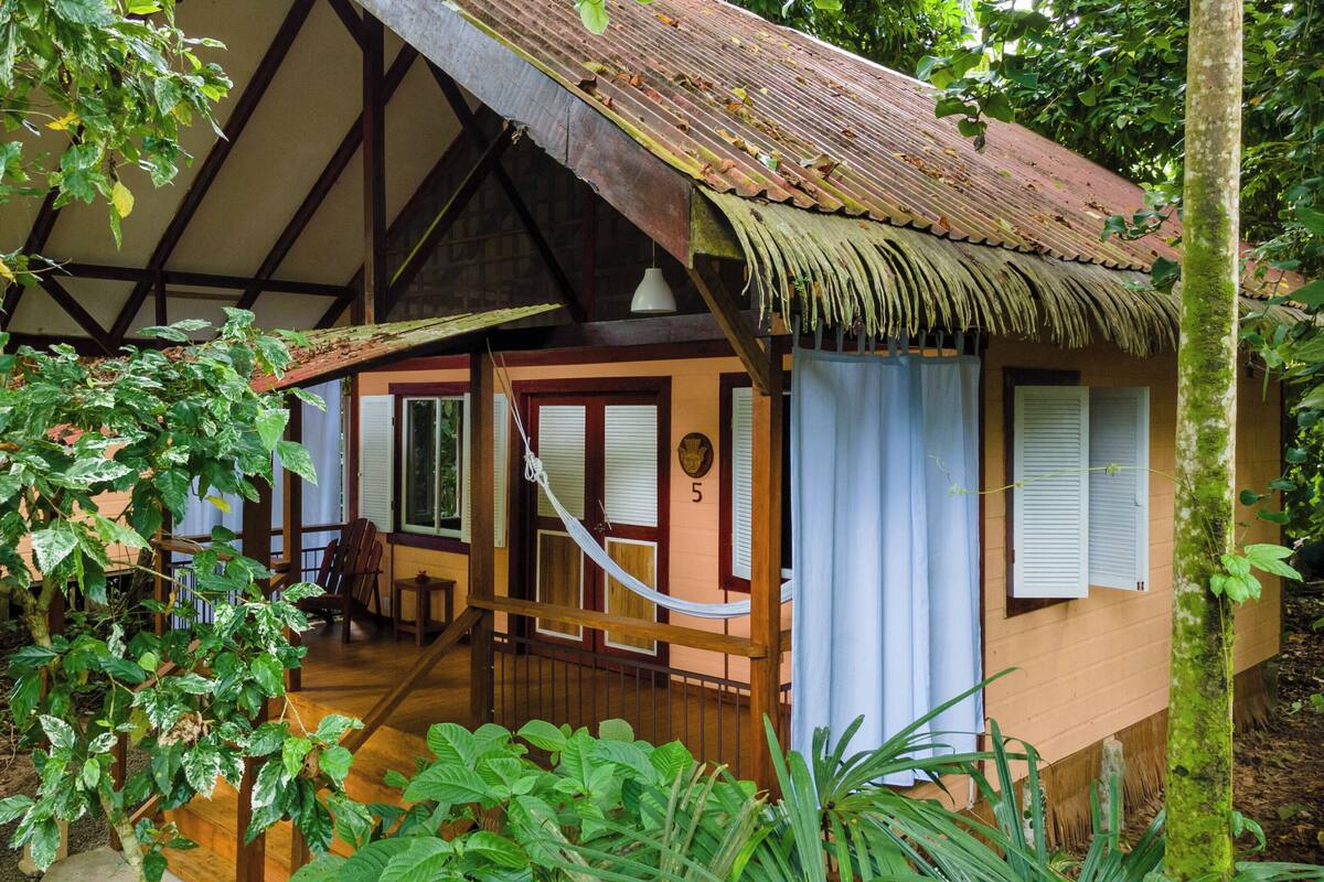 A rustic wooden house with a thatched roof, a wooden porch, and a large window.