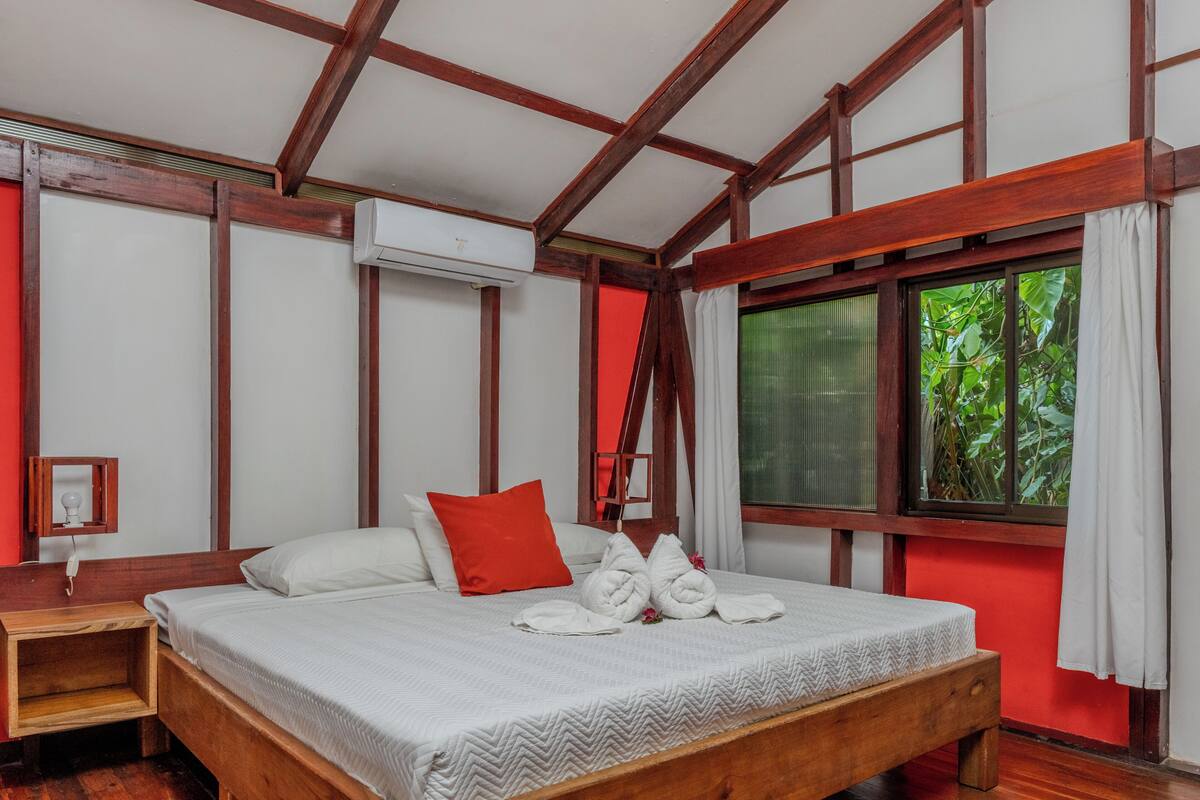 A room with a wooden bed, a red accent wall, an air conditioning unit, a window with a view of greenery, and a bedside table with a lamp.