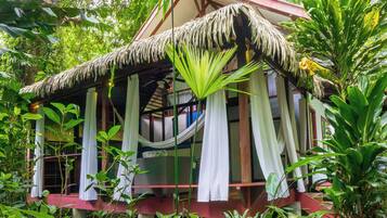 A thatched-roof structure surrounded by lush greenery, with white curtains and a hammock hanging from the ceiling.