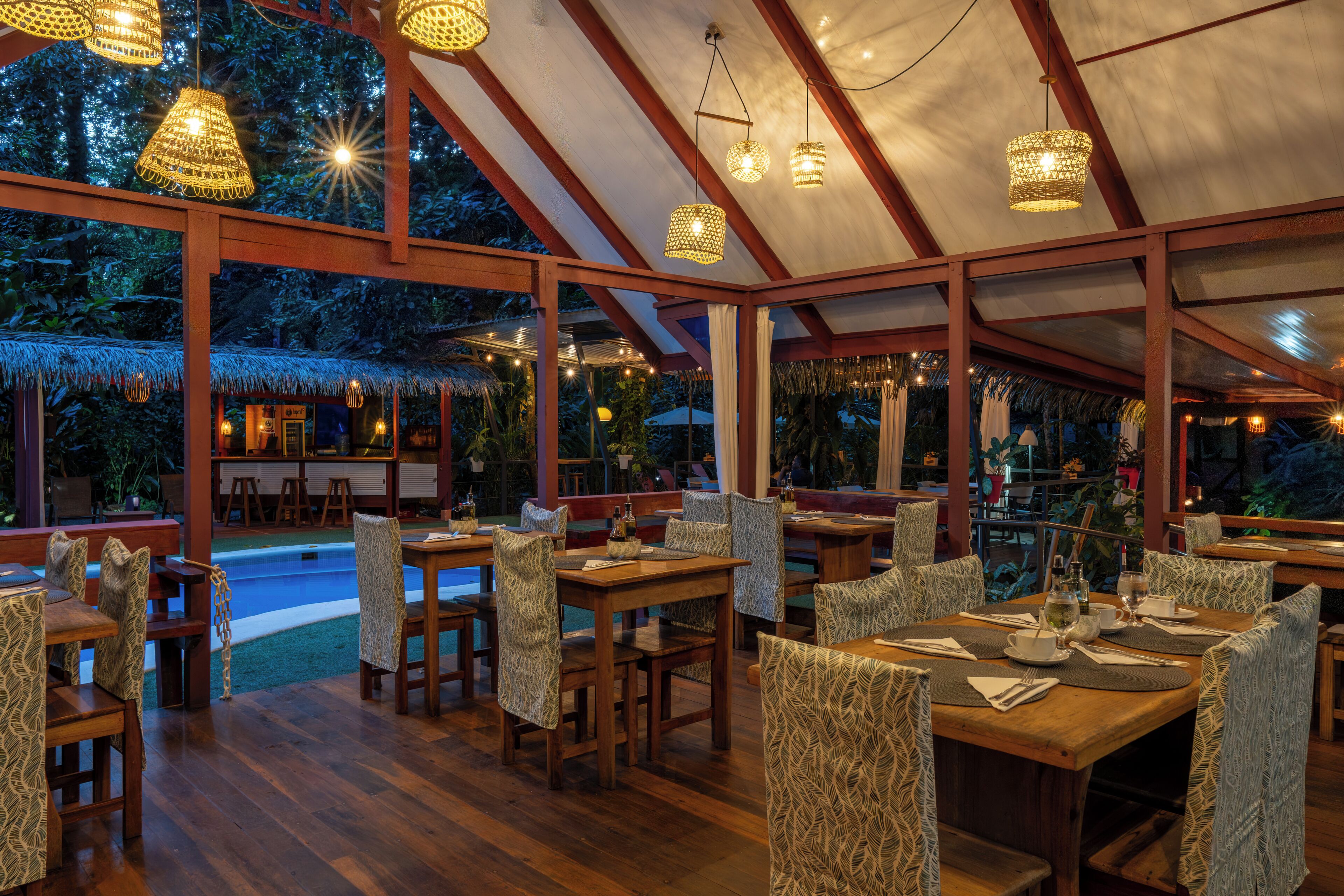 Un comedor interior con mesas y sillas de madera, una piscina y una estructura de techo de paja.