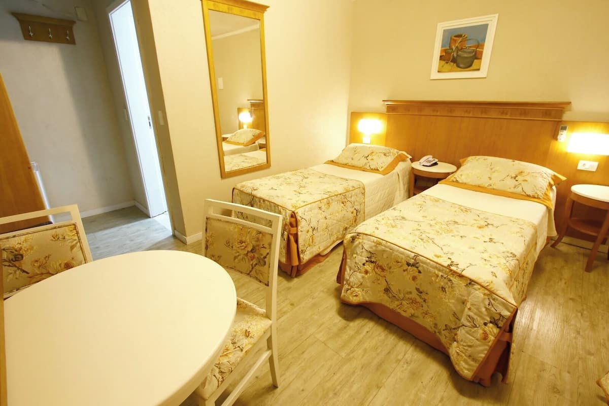 A hotel room with two single beds, a round table, a chair, a mirror, and a bedside table with a lamp.