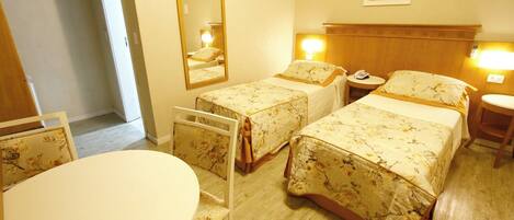 Habitación de hotel con dos camas individuales, una mesa redonda, una silla, un espejo y una mesita de noche con lámpara.
