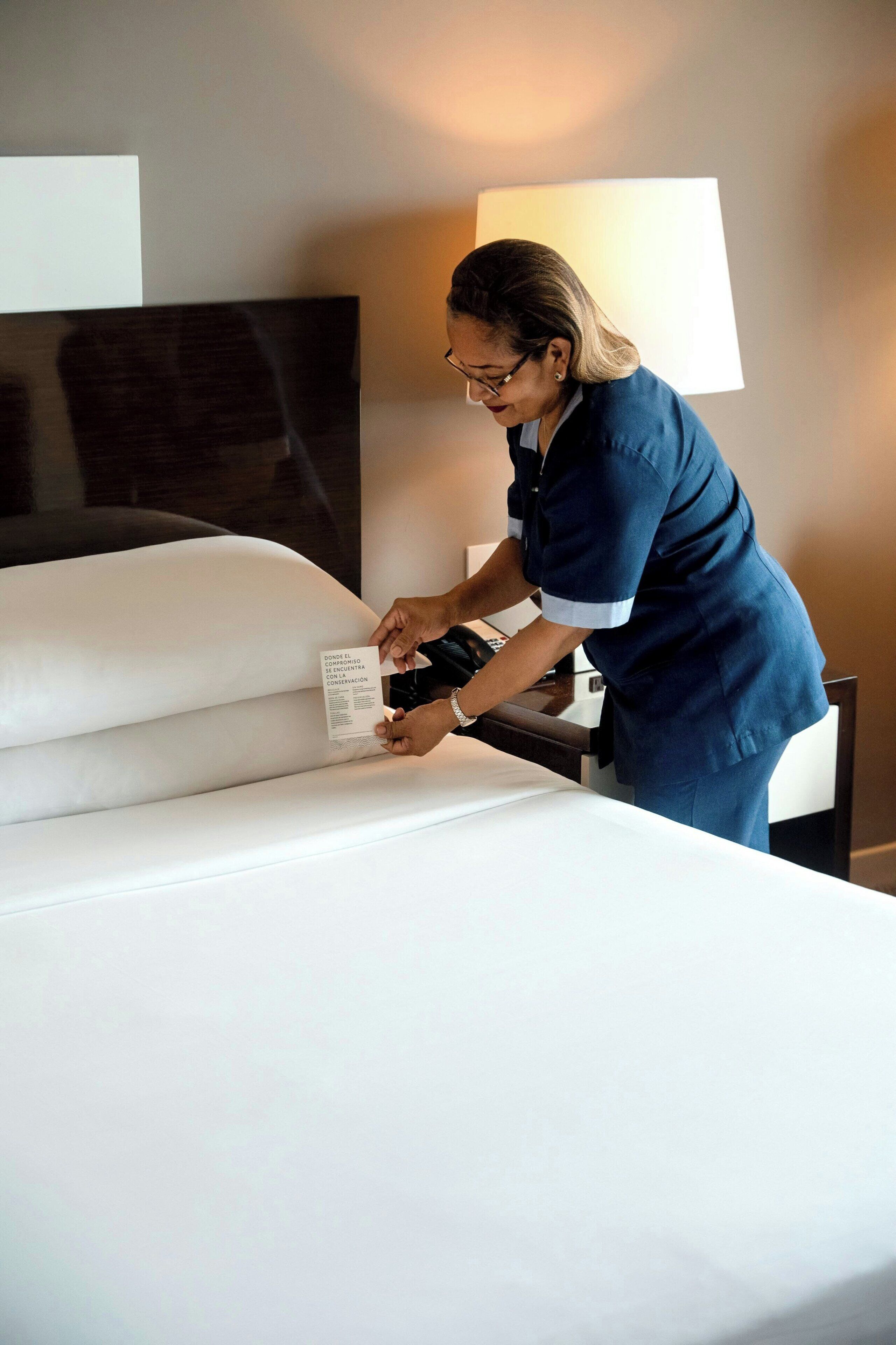 A person in a blue uniform adjusting a white bedspread in a hotel room with a large window and a flat-screen TV on the wall.