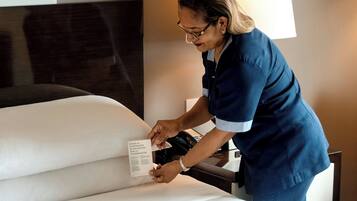 A person in a blue uniform adjusting a white bedspread in a hotel room with a large window and a flat-screen TV on the wall.