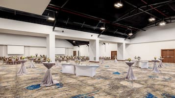 Um salão amplo com mesas e cadeiras brancas, um palco e um teto alto com vigas aparentes.