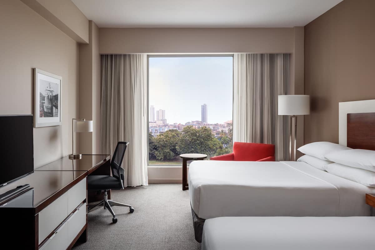 A hotel room with a bed, a desk with a chair, a TV, and a view of the city through large windows.