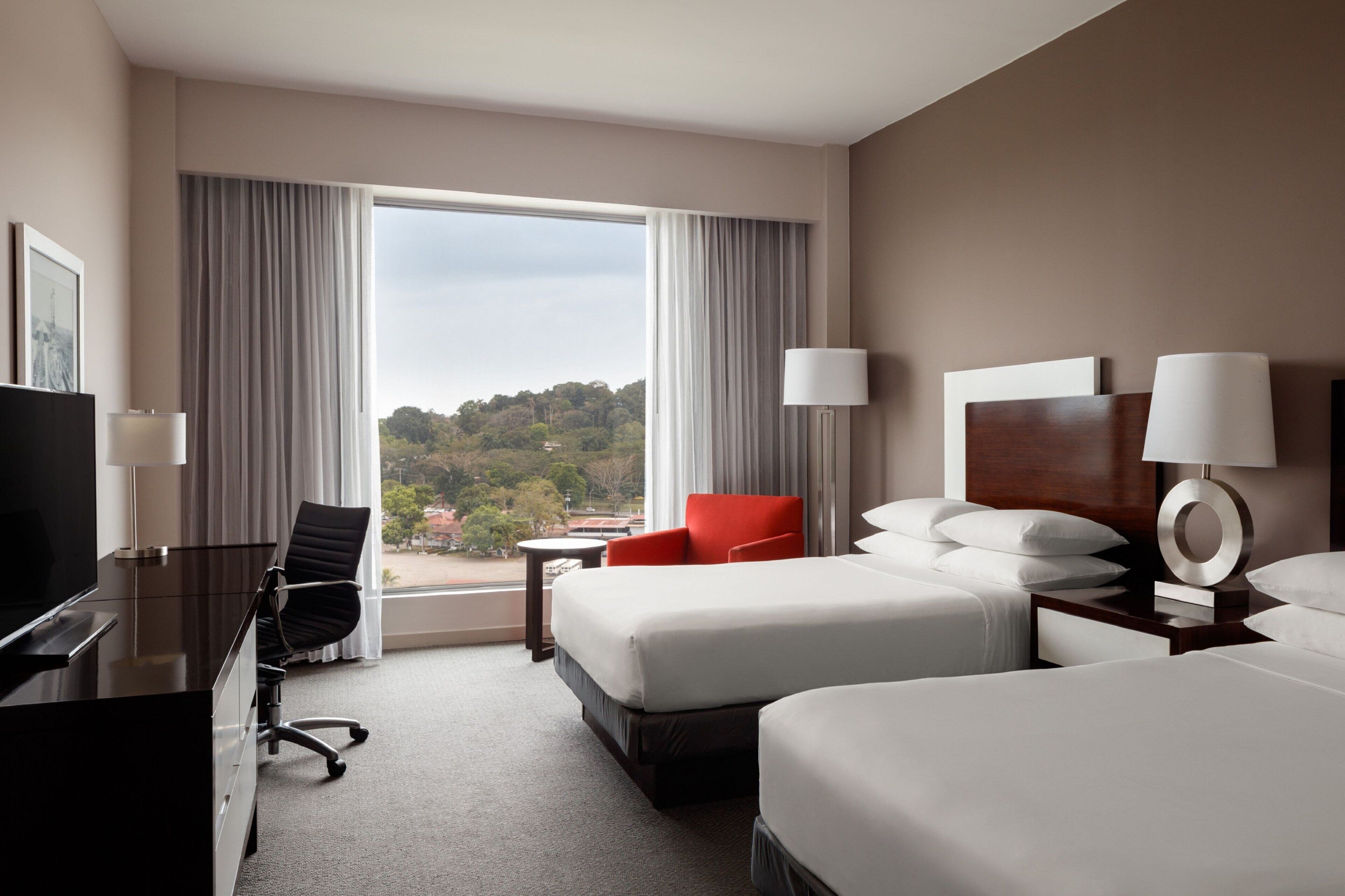 A hotel room with two beds, a desk, a chair, and a TV. There is a window with curtains and a view of trees.