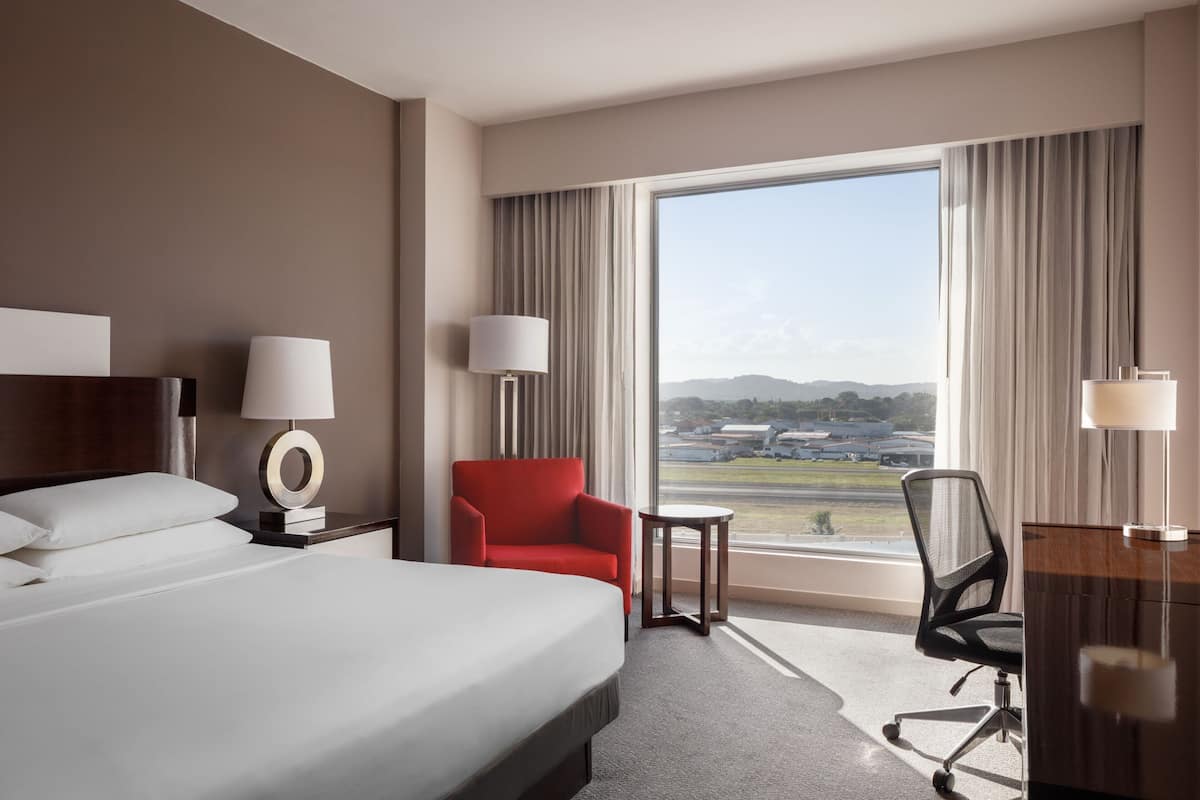 A hotel room with a large bed, a desk, a chair, a small table, and a view of an airport.