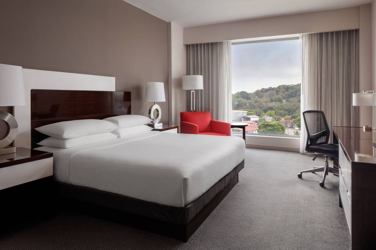 A hotel room with a large bed, two bedside tables with lamps, a red chair, a desk with a chair, and a large window with a view of the city.