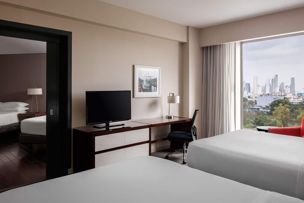A hotel room with a bed, a desk with a TV, a chair, and a large window offering a city view.