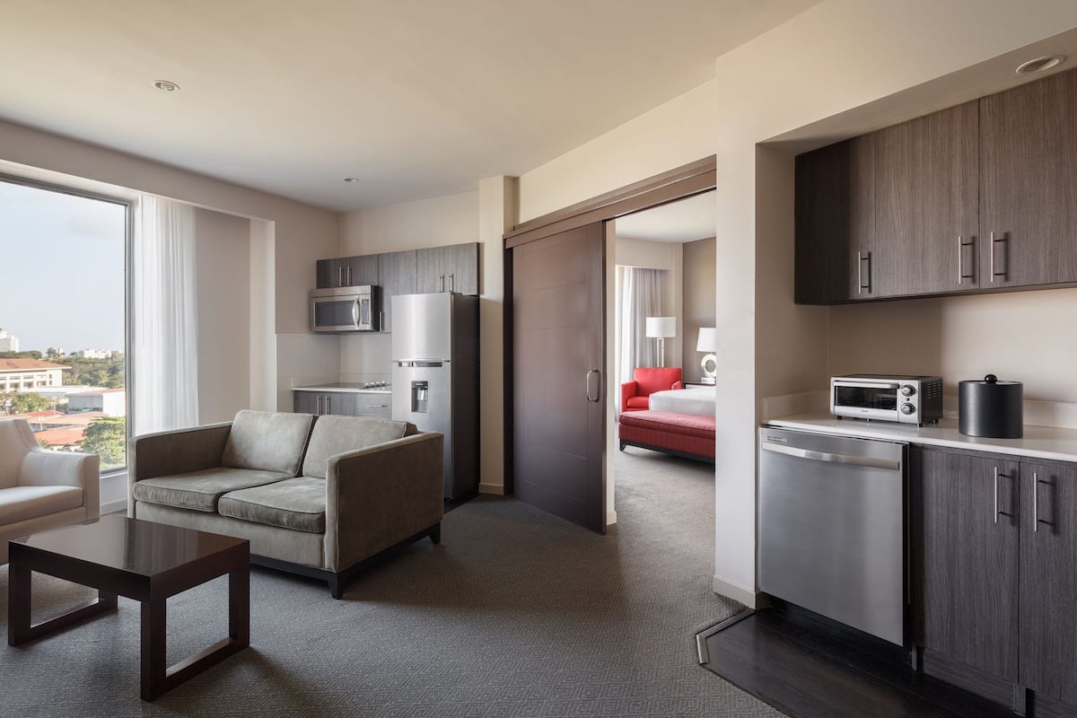 A modern hotel room with a sofa, a small table, a kitchen area with a microwave, refrigerator, and a dishwasher.